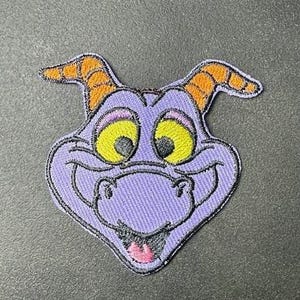 Könnte beinhalten: Gestickter Aufnäher mit einem lila Cartoon-Charakter mit orangefarbenen Hörnern, gelben Augen und einer rosa Zunge. Der Aufnäher hat eine schwarze Umrandung, für Kleidung oder Accessoires.