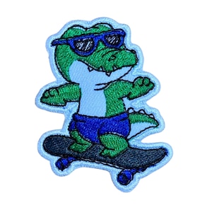 Op de afbeelding: Geborduurde patch met een groene alligator met een zonnebril en een blauwe korte broek, skateboardend op een zwart skateboard. De patch heeft een lichtblauwe rand, geschikt voor kleding of accessoires.