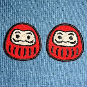 May include: Two red Daruma doll patches with black outlines and white facial features are displayed on a blue denim background. The Daruma dolls have a traditional Japanese design, with a round shape and stylized eyes.