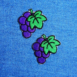 Può includere: Due toppe ricamate raffiguranti un grappolo di uva viola con foglie verdi. Le toppe hanno un bordo bianco e sono poste su uno sfondo di tessuto blu. Ideali per decorare abbigliamento o accessori.