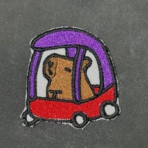 Peut inclure: Écusson brodé représentant un ours brun conduisant une voiture rouge avec un toit violet. L'ours est visible à travers les fenêtres de la voiture. L'écusson a une bordure blanche et des roues noires.