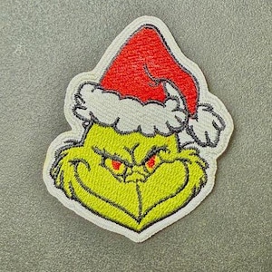 Può includere: Toppa ricamata raffigurante un personaggio verde con un cappello da Babbo Natale rosso con bordi bianchi. Il personaggio ha un'espressione accigliata con occhi rossi. La toppa è bordata di bianco, per abbigliamento o accessori.