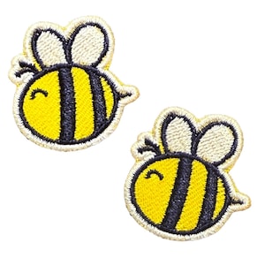 Könnte beinhalten: Zwei gestickte Bienenaufnäher. Jeder Aufnäher hat einen gelben Körper mit schwarzen Streifen, weiße Flügel und ein lächelndes Gesicht. Die Aufnäher sind mit einem gestickten Rand versehen.