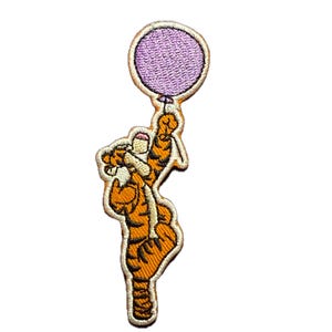 Op de afbeelding: Geborduurde patch met Tijgertje uit Winnie de Poeh, met een paarse ballon. De patch heeft oranje en zwarte strepen, met een witte rand. De ballon is lichtpaars.