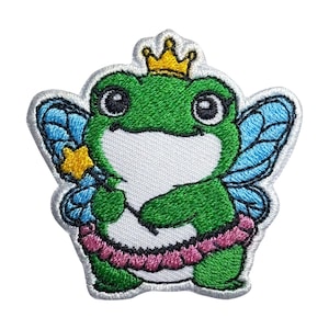 Puede incluir: Parche bordado para planchar de una rana verde con una corona dorada y alas de hada azules, sosteniendo una varita con una estrella amarilla. La rana lleva un tutú rosa.