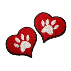 May include: Two red heart-shaped patches with a white paw print design. Each patch has a black border. The patches are made of fabric and are likely embroidered. The patches are designed for pet lovers.