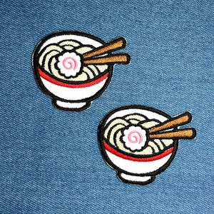 Peut inclure: Deux écussons brodés représentant des bols de ramen. Chaque bol blanc est orné d'une bande rouge et d'une bordure noire, rempli de nouilles, d'une spirale rose et de baguettes marron. Sur fond de denim bleu.