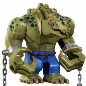 May include: A Lego figure of Killer Croc, a DC Comics villain. The figure is mainly olive green with a crocodile-like head and torso, blue trousers, and silver shackles. Chains are attached to the figure's wrists.