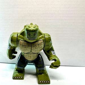 Peut inclure: Figurine d'action verte et beige représentant un personnage humanoïde reptilien. La figurine a un visage détaillé avec des dents pointues et un corps texturé. La figurine porte un pantalon noir et des rayures blanches sur les bras.