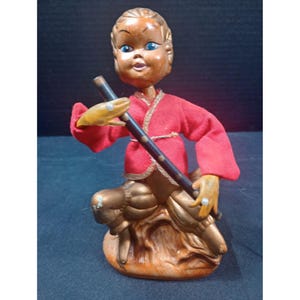Könnte beinhalten: Vintage Figur einer sitzenden Figur in roter Robe, die eine dunkle Flöte hält. Die Figur hat einen Holzkopf mit blauen Augen und sitzt auf einem braunen Sockel. Die Hände und Beine der Figur sind goldfarben bemalt.