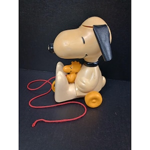 Op de afbeelding: Vintage Snoopy trekfiguur met Woodstock. Het speelgoed heeft een beige Snoopy-figuur met zwarte oren en kraag, die een gele Woodstock vasthoudt. Het heeft een rood trekkoord en gele wielen. Een klassiek speelgoed.