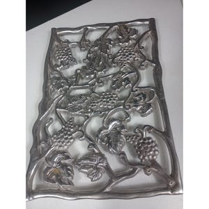 Puede incluir: Un salvamanteles rectangular de metal plateado con un diseño de vid. El salvamanteles presenta representaciones detalladas de racimos de uvas y hojas, creando un accesorio de cocina decorativo y funcional.