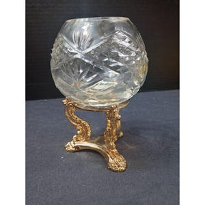 May include: A clear, round crystal bowl with a cut-glass pattern, resting on an ornate gold-colored metal stand. The stand has a three-legged base and decorative scrollwork. The bowl is likely for decorative purposes.