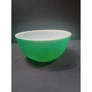 Op de afbeelding: Een groene glazen kom met een witte binnenkant. De kom heeft een gladde, afgeronde vorm en een glanzende afwerking. De buitenkant is een levendige groene kleur, terwijl de binnenkant wit is. De kom is leeg en lijkt in goede staat te verkeren.