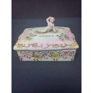 May include: A rectangular porcelain trinket box with a lid, featuring a reclining cherub figurine on top. The box is decorated with gold floral patterns and a pastel color scheme of pink, yellow, and white. The box has a decorative border.