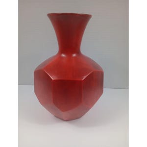 Puede incluir: Un jarrón rojo mate con un cuerpo geométrico de múltiples facetas y un cuello liso y acampanado. El recipiente presenta una estructura hexagonal distintiva con paneles planos que se estrechan hacia la base y se extienden hacia arriba.