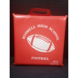 Puede incluir: Un cojín de asiento cuadrado de vinilo rojo con un asa integrada. El frente presenta un gráfico blanco de un balón de fútbol americano y el texto "ROSWELL HIGH SCHOOL FOOTBALL" impreso en blanco. Un pequeño logo es visible abajo.