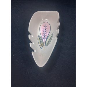 May include: A light grey, leaf-shaped ceramic dish with a pink and green floral design. The dish has a scalloped edge and is likely used for serving small items or as a decorative piece. The floral design features a pink leaf with green and blue accents.