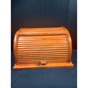 Pode incluir: Uma caixa de pão retangular de madeira com um acabamento liso em castanho claro. O recipiente apresenta uma porta de correr curva com ripas que desliza para cima, equipada com um pequeno puxador de madeira centrado na ripa inferior.