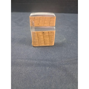 Puede incluir: Un tapón de botella rectangular de dos partes, con una sección superior e inferior transparente. La sección central es de color marrón claro con un patrón texturizado en forma de rejilla. El tapón está diseñado para encajar en una botella.