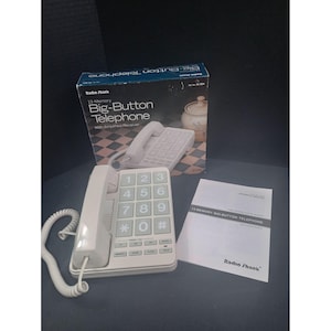 May include: A white corded telephone with large, light grey buttons, a handset, and a coiled cord. The set includes the original box labelled 13-Memory Big-Button Telephone with Amplified Receiver and a printed owner's manual from Radio Shack.