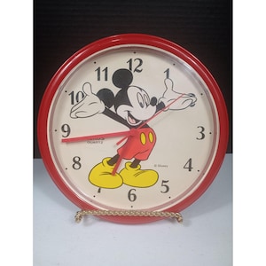 May include: A red-rimmed quartz clock featuring Mickey Mouse. The clock face is cream-colored with black numerals and red hands. Mickey Mouse is depicted in his classic outfit, with yellow shoes and red shorts. The clock is on a gold-colored stand.