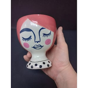 May include: A ceramic face planter with a pink head, blue eyes, and pink cheeks. The white face has a black dotted collar. The planter is held in a hand.