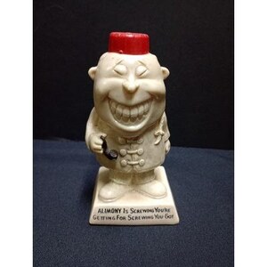 May include: A vintage figurine of a smiling man wearing a red fez and holding a pipe. The figurine is off-white with a detailed face and clothing. The base reads "ALIMONY IS SCREWING YOU'RE GETTING FOR SCREWING YOU GOT."