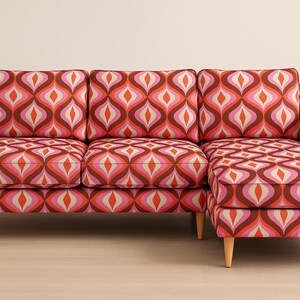 Könnte beinhalten: Ein Ecksofa im Retro-Stil mit einem lebendigen geometrischen Muster in Rosa-, Orange- und Brauntönen. Das Sofa hat Holzbeine und eine Chaiselongue auf einer Seite und bietet eine stilvolle Sitzgelegenheit.