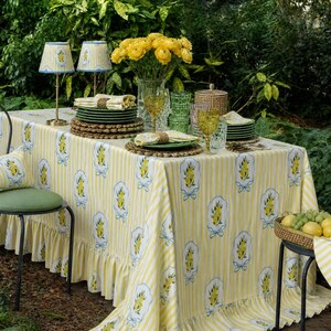 Puede incluir: Una mesa de comedor al aire libre bellamente decorada con un mantel a rayas amarillas y blancas con motivos de limones. La mesa está adornada con platos verdes, copas doradas y un jarrón con flores amarillas, creando un ambiente acogedor.
