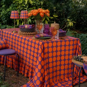 May include: A vibrant outdoor dining scene featuring a table covered in an orange and purple polka dot tablecloth. The table is set with plates, glasses, and napkins. Two lamps with matching shades and a vase of orange flowers add to the decor.
