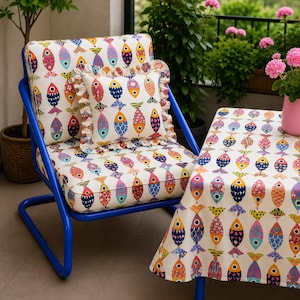May include: A bright blue metal chair with a white cushion featuring a colorful fish pattern. A matching square throw pillow with a ruffled edge sits on the seat. A table covered in a matching fish-patterned tablecloth stands next to the chair.