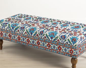 May include: A rectangular ottoman with a white background and a floral pattern in red and blue. The ottoman has wooden legs and a decorative border. The floral pattern is repeated across the top and sides.