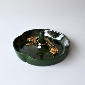 May include: A dark green ceramic ring dish with a central post for rings. A gold necklace and a ring are displayed on the dish. The dish has a unique, organic shape and a glossy finish.