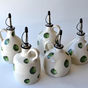 May include: A set of four cream-coloured ceramic oil dispensers with rounded bodies, side handles, and silver-toned metal pour spouts with black caps. Each dispenser features a hand-painted pattern of scattered green circular spots across the surface.