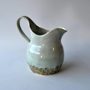 May include: A ceramic pitcher with a glossy light blue-grey glaze on the upper portion and a textured, honeycomb-like pattern in a muted ochre tone at the base. The vessel features a curved handle and a distinct pouring spout with a smooth finish.
