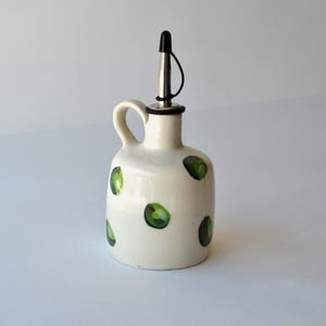 May include: A cream-coloured ceramic oil cruet with a rounded body and a single handle. The exterior features hand-painted green circular spots. A stainless steel pour spout with a black plastic cap is inserted into the narrow neck of the vessel.