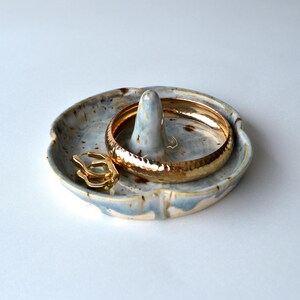 May include: A ceramic ring dish with a central post and a shallow bowl. The dish is glazed in shades of blue, grey, and gold. A gold bracelet and ring rest on the dish, showcasing jewelry storage.
