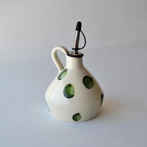 May include: A cream-coloured ceramic oil cruet with a wide base and a single handle. The exterior features hand-painted green olive motifs scattered across the surface. A stainless steel pourer with a black plastic cap is inserted into the top.