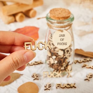 Puede incluir: Un tarro de cristal transparente con tapa de corcho, etiquetado "JAR OF FUCKS", lleno de recortes de madera. La palabra "FUCK" está escrita con letras de madera, algunas esparcidas alrededor del tarro. Una mano sostiene un recorte de madera "FUCK".