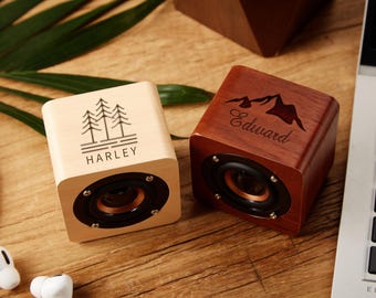 Può includere: Due piccoli altoparlanti Bluetooth in legno con incisioni personalizzate. Un altoparlante è di colore marrone chiaro con un design di tre alberi e il nome "HARLEY". L'altro altoparlante è di colore marrone scuro con un design di montagna e il nome "Edward".