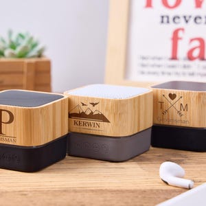 May include: Three small, square, wooden Bluetooth speakers with different designs. The speaker on the left has a black and brown finish with the name "Patrick" and the letter "P" engraved on it. The speaker in the middle has a grey and brown finish with the name "Kerwin" and a mountain range design engraved on it. The speaker on the right has a brown finish with the letters "T" and "M" and a heart design engraved on it. The text "Groomsman" is engraved on all three speakers.