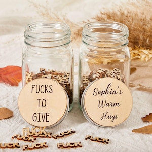 Può includere: Due barattoli di vetro con coperchi in legno. Un coperchio recita "Fucks To Give", l'altro "Sophia's Warm Hugs". I barattoli contengono forme di lettere di legno. Lettere di legno sparse compongono "FUCK".