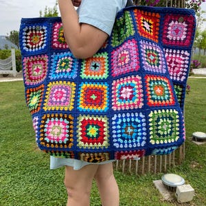 May include: A large, handmade tote bag with a colorful patchwork design. The bag features a dark blue border and is constructed from numerous crocheted squares in various colors, including pink, yellow, green, and orange. It has a black strap.
