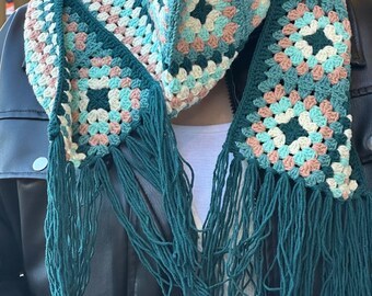 May include: A green crocheted scarf with fringe, featuring a geometric pattern in shades of pink, blue, and white.