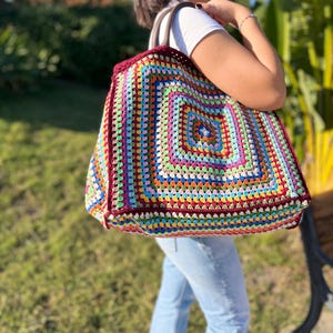 Może przedstawiać: Duża, kolorowa torba na ramię wykonana na szydełku z kwadratowym wzorem w odcieniach czerwieni, zieleni, błękitu, żółci i bieli. Torba ma brązowy skórzany uchwyt.