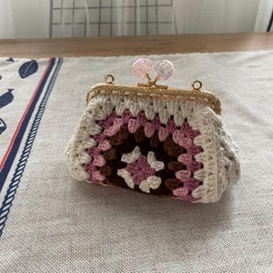 Op de afbeelding: Een gehaakte portemonnee in een granny square patroon, met witte, roze en bruine vierkanten. Het heeft een goudkleurig metalen frame met een decoratieve sluiting en twee iriserende kraalaccenten.