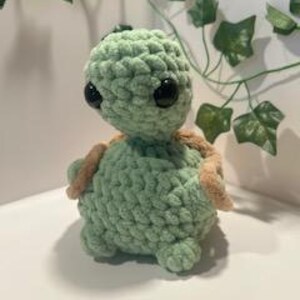 May include: A handmade, light green crochet turtle with a textured shell and large black eyes. The turtle has tan-colored flippers and a small head. The background includes artificial ivy.