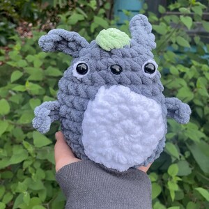 May include: A grey crocheted plush toy with large black eyes and a green leaf on top. The toy is being held by a hand.