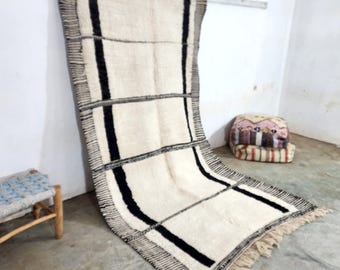 May include: A white and black Moroccan rug with a geometric pattern. The rug has a fringe border and is made of wool.